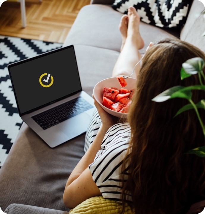 Femeie întinsă pe o canapea mâncând un bol de fructe cu un laptop lângă ea afișând logo-ul Norton